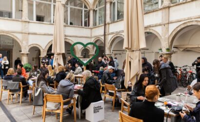 Viele Menschen sitzen an Holztischen im Innenhof eines historischen Gebäudes und frühstücken gemeinsam. In der Mitte steht ein großes, grünes Herz mit der Aufschrift „GAMLITZ“. Auf einem Hocker steht „Creative Industries Styria“. Die Atmosphäre wirkt lebendig und einladend.