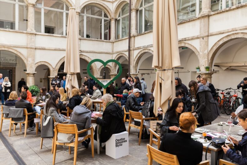 Viele Menschen sitzen an Holztischen im Innenhof eines historischen Gebäudes und frühstücken gemeinsam. In der Mitte steht ein großes, grünes Herz mit der Aufschrift „GAMLITZ“. Auf einem Hocker steht „Creative Industries Styria“. Die Atmosphäre wirkt lebendig und einladend.