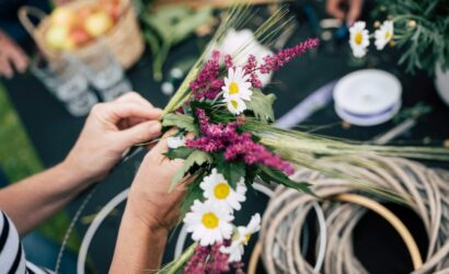 Hände flechten einen dekorativen Kranz aus Getreidehalmen, weißen Margeriten und violetten Blüten auf einem Tisch mit Bastelmaterialien.