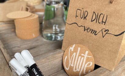 Im Vordergrund steht eine braune Geschenktüte aus Kraftpapier mit der Aufschrift "Für dich vom ♥". Daneben lehnt ein runder Korkuntersetzer mit dem handgeschriebenen Wort "Salute" in stilvollem Weiß. Drei Kreidemarker liegen in einer hölzernen Ablage. Im Hintergrund befinden sich Bastelmaterialien wie Korkscheiben, ein Glas Wasser und ein geflochtener Korb mit getrocknetem Lavendel.