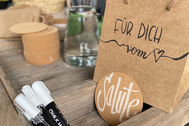Im Vordergrund steht eine braune Geschenktüte aus Kraftpapier mit der Aufschrift "Für dich vom ♥". Daneben lehnt ein runder Korkuntersetzer mit dem handgeschriebenen Wort "Salute" in stilvollem Weiß. Drei Kreidemarker liegen in einer hölzernen Ablage. Im Hintergrund befinden sich Bastelmaterialien wie Korkscheiben, ein Glas Wasser und ein geflochtener Korb mit getrocknetem Lavendel.