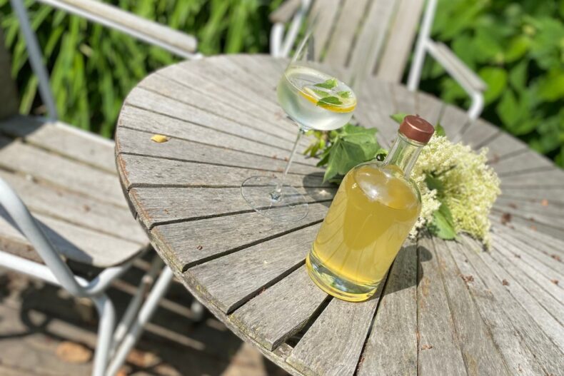 Ein runder Holztisch mit wettergegerbter Oberfläche steht auf einer Terrasse im Grünen. Darauf befinden sich eine Glasflasche mit gelblichem Sirup, ein Glas mit einem erfrischenden Getränk, garniert mit Zitronenscheibe und Minze, sowie ein kleiner Strauß Holunderblüten. Im Hintergrund sind zwei Metallstühle zu sehen.