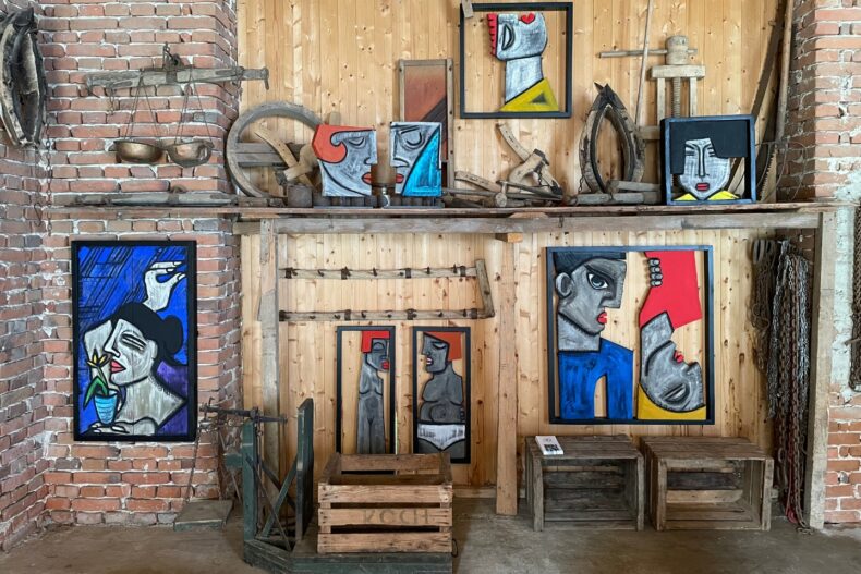 Eine rustikale Wand aus Backstein und Holz dient als Kulisse für eine Kunstausstellung. Mehrere abstrakte Gemälde mit kantigen, geometrischen Gesichtern in kräftigen Farben wie Blau, Rot und Gelb sind ausgestellt. Umgeben werden die Kunstwerke von alten landwirtschaftlichen Werkzeugen, Waagen und Holzkisten.
