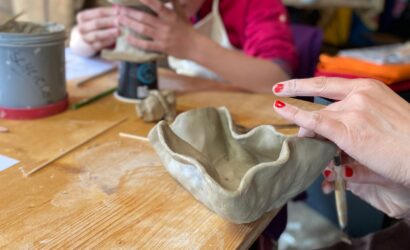Zwei Personen arbeiten an Tonobjekten in einer Keramikwerkstatt; im Vordergrund wird eine handgeformte Schale bearbeitet.