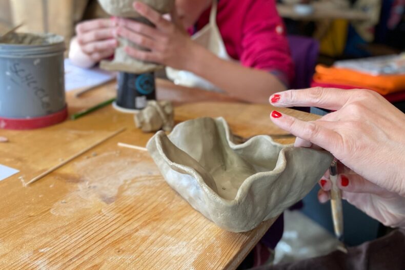 Zwei Personen arbeiten an Tonobjekten in einer Keramikwerkstatt; im Vordergrund wird eine handgeformte Schale bearbeitet.
