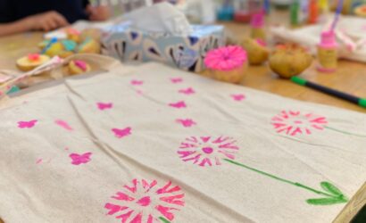 Ein bemalter Stoffbeutel mit pinken Blumen- und Schmetterlingsmotiven sowie grünen Stielen und Blättern liegt auf einem Holztisch.