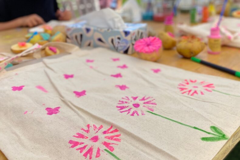 Ein bemalter Stoffbeutel mit pinken Blumen- und Schmetterlingsmotiven sowie grünen Stielen und Blättern liegt auf einem Holztisch.