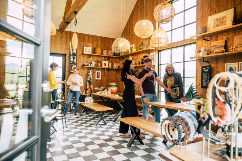 Das Bild zeigt einen stilvoll eingerichteten Hofladen mit handgefertigten Produkten und kunstvollen Accessoires. Die Atmosphäre ist warm und einladend, mit Holzwänden, großen Fenstern und dekorativen Lampen. Mehrere Personen sind in Gespräche vertieft und betrachten die ausgestellten Waren.