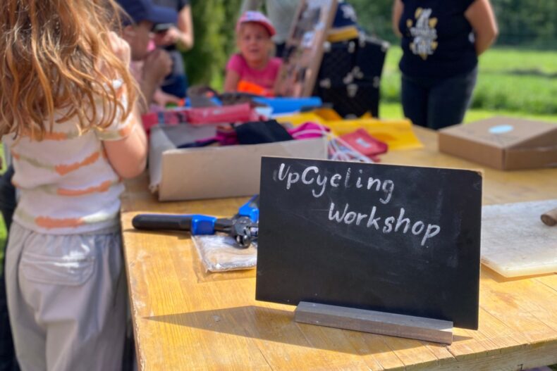 Ein Holztisch im Freien mit einer kleinen Tafel, auf der „UpCycling Workshop“ steht. Auf dem Tisch liegen verschiedene Materialien und Werkzeuge. Im Hintergrund sind Kinder und Erwachsene unter einem Sonnenschirm zu sehen.
