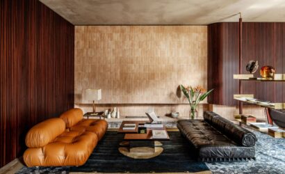 Modernes Wohnzimmer mit einem braunen Ledersofa, einem schwarzen Ledertagesbett, einem Holztisch und dekorativen Objekten. Die Wände sind mit Holzpaneelen und beigen Fliesen gestaltet, dazu ein dunkler Teppich mit geometrischem Muster.
