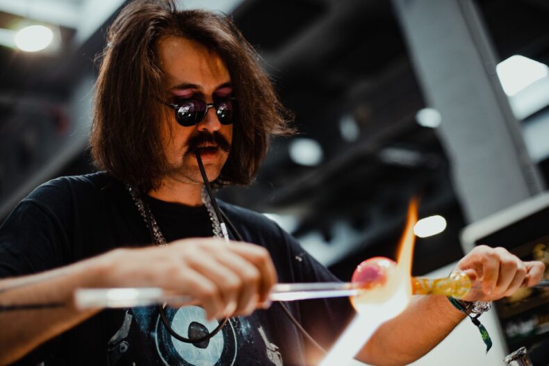 Ein Glasbläser mit langen Haaren, Schnurrbart und runden Schutzbrillen arbeitet konzentriert an einem glühenden Glasstück. Er hält Glasstäbe in beiden Händen und formt das Material mit Hilfe einer offenen Flamme. Im Hintergrund ist eine industrielle Umgebung mit unscharfen Lichtern zu erkennen.