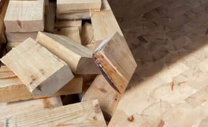 Stapel grob geschnittener Holzstücke neben einem glatt verlegten Holzpflasterboden aus quadratischen Holzblöcken mit sichtbarer Jahresringstruktur. Das Bild zeigt den Kontrast zwischen rohem und verarbeitetem Holz.