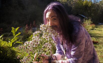 Eine Person mit langen, lila Haaren und einem violetten Gewand riecht an einem Strauß violett-weißer Wildblumen. Sie trägt zahlreiche Armbänder und Anhänger. Im Hintergrund ist ein grüner Garten mit Pflanzen, Sträuchern und einem kleinen Gebäude zu sehen.