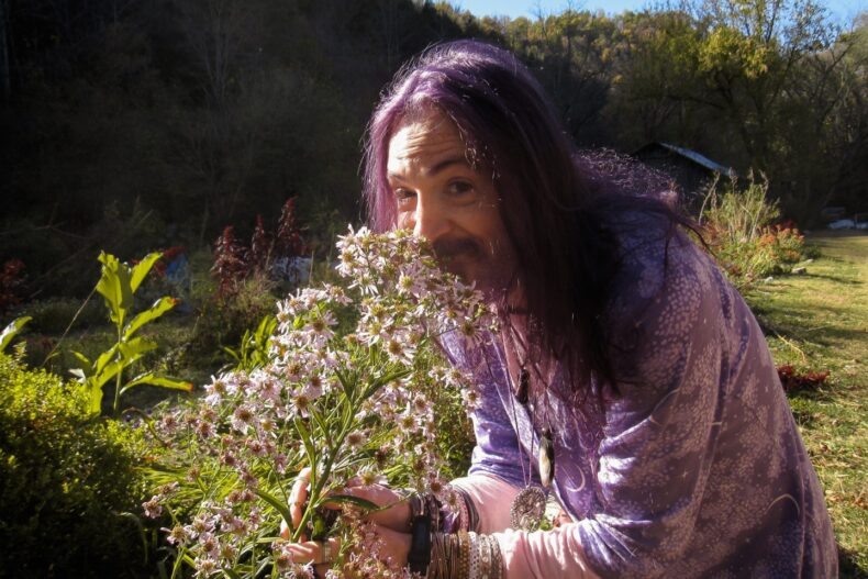Eine Person mit langen, lila Haaren und einem violetten Gewand riecht an einem Strauß violett-weißer Wildblumen. Sie trägt zahlreiche Armbänder und Anhänger. Im Hintergrund ist ein grüner Garten mit Pflanzen, Sträuchern und einem kleinen Gebäude zu sehen.