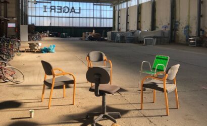 Fünf verschiedene Stühle, darunter ein Bürostuhl und ein grüner Gartenstuhl, stehen kreisförmig in einer leerstehenden Industriehalle. Im Hintergrund sind Fahrräder, Paletten und abgedeckte Möbel zu sehen. Das Wort „LAGER1“ ist spiegelverkehrt an den Fenstern zu lesen.