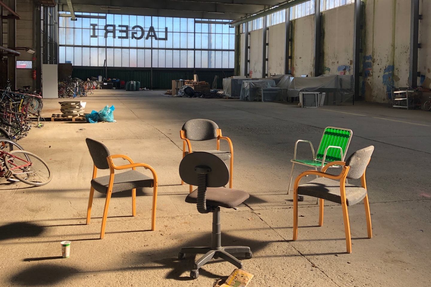 Fünf verschiedene Stühle, darunter ein Bürostuhl und ein grüner Gartenstuhl, stehen kreisförmig in einer leerstehenden Industriehalle. Im Hintergrund sind Fahrräder, Paletten und abgedeckte Möbel zu sehen. Das Wort „LAGER1“ ist spiegelverkehrt an den Fenstern zu lesen.