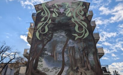 Großes Wandgemälde an einer Hausfassade mit einer Waldszene, in der eine Person mit zwei Hunden spazieren geht. Über dem Wald sind abstrakte, grüne Muster und ein lila Stern zu sehen.