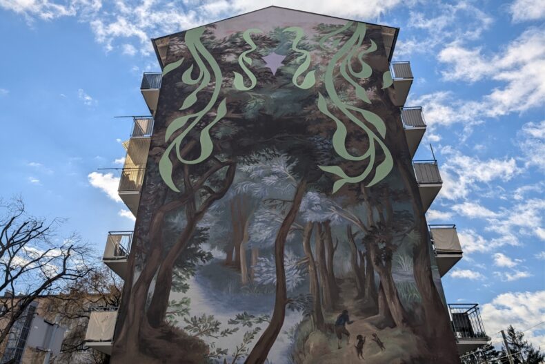 Großes Wandgemälde an einer Hausfassade mit einer Waldszene, in der eine Person mit zwei Hunden spazieren geht. Über dem Wald sind abstrakte, grüne Muster und ein lila Stern zu sehen.