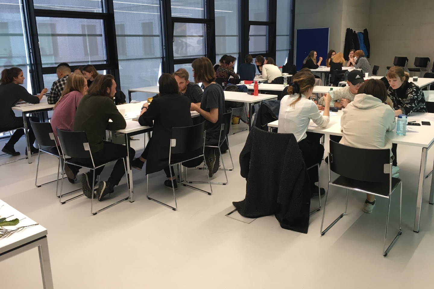 Mehrere Gruppen von Menschen sitzen an weißen Tischen in einem modernen Seminarraum mit großen Fenstern. Sie diskutieren, schreiben und arbeiten gemeinsam an Projekten. Die Atmosphäre wirkt konzentriert und kollaborativ.
