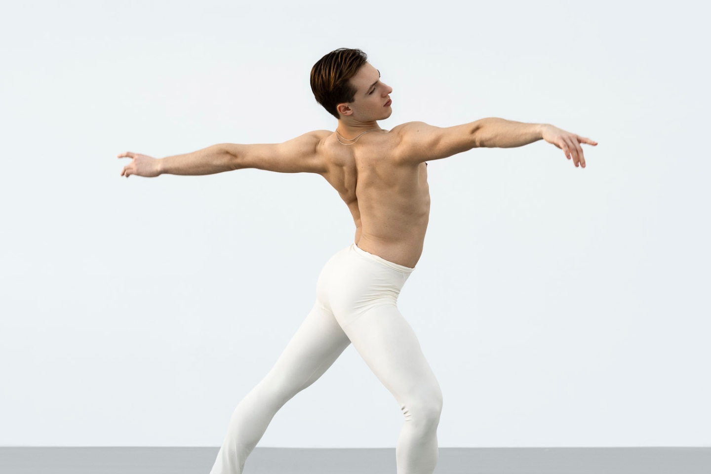 Ein Balletttänzer mit athletischem, muskulösem Körper bewegt sich anmutig in einer klassischen Pose. Er trägt eine eng anliegende, weiße Ballettstrumpfhose und ist oberkörperfrei. Der Hintergrund ist minimalistisch und hell.