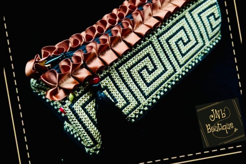 Handgefertigte Wayuu-Tasche mit schwarz-beigem, geometrischem Muster und dekorativer, kupferfarbener Rüschenverzierung am Rand. Kleine Perlen und Quasten ergänzen das Design. Das Logo „JNB Boutique“ ist in der Ecke sichtbar.