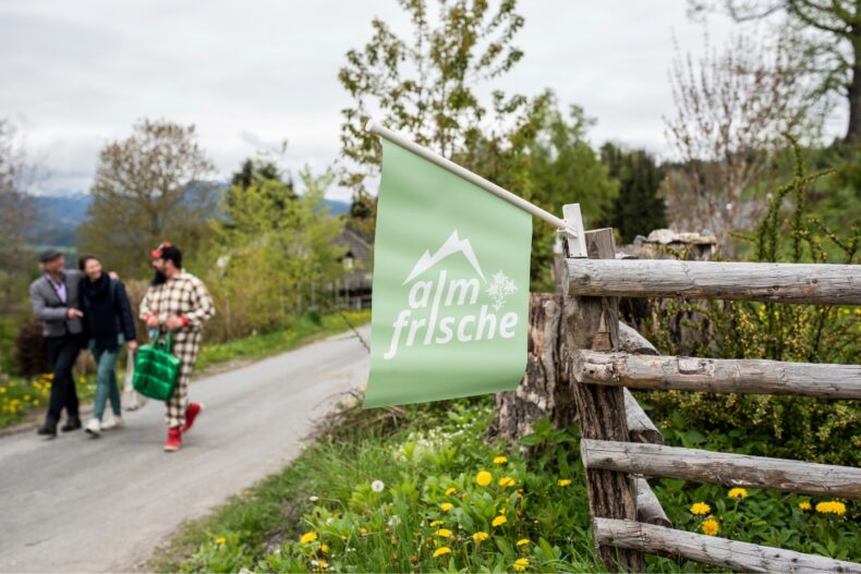 Eine grüne Fahne mit der Aufschrift „almfrische“ und einem stilisierten Berglogo weht an einem rustikalen Holzzaun. Im Hintergrund spazieren drei Personen eine ländliche Straße entlang, umgeben von grüner Natur und blühendem Löwenzahn.