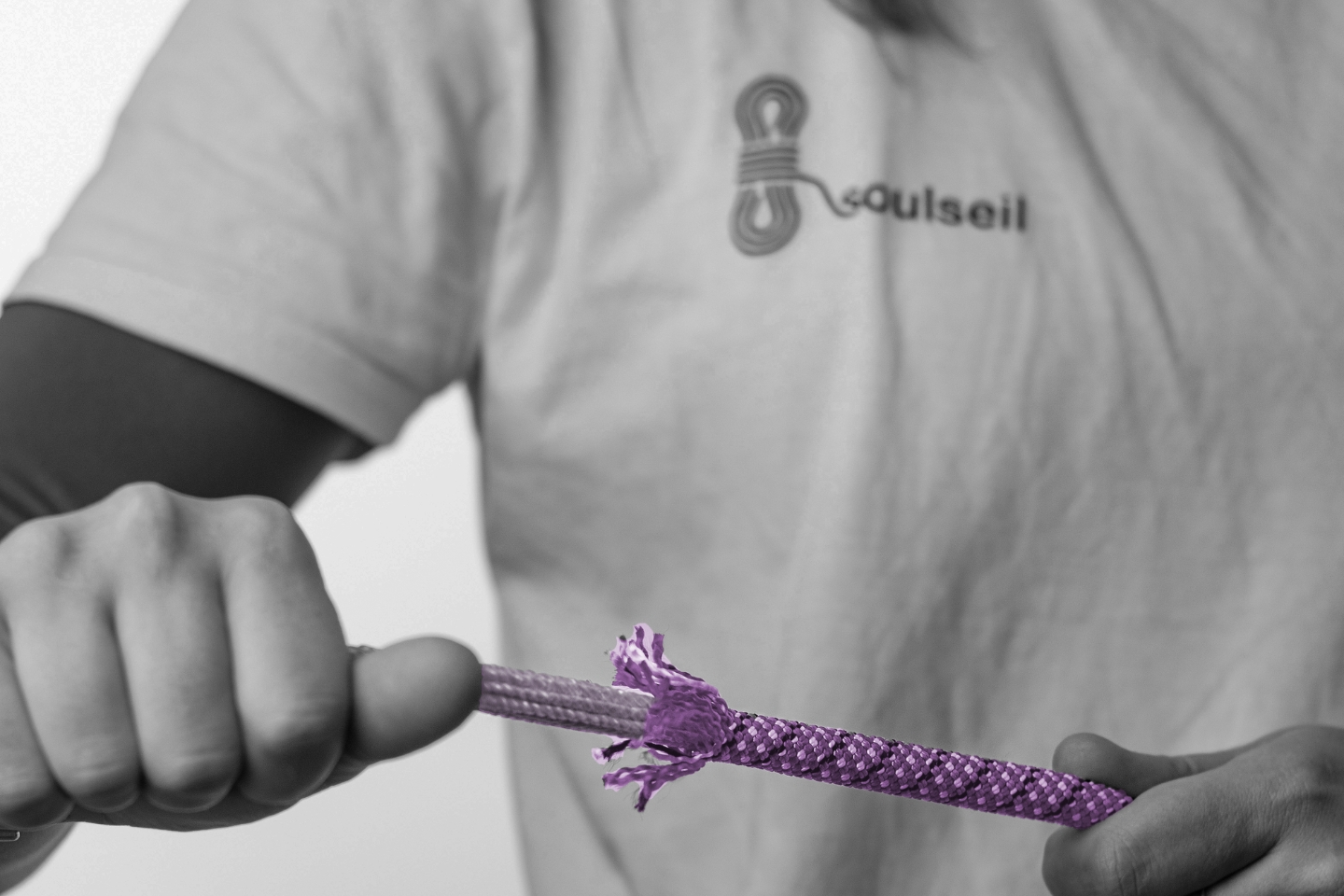 Eine Person im T-Shirt mit der Aufschrift „soulseil“ hält ein ausgefranstes, lilafarbenes Kletterseil in den Händen. Der Hintergrund sowie die Kleidung sind in Schwarz-Weiß gehalten, während nur das Seil farbig hervorgehoben ist. Das Bild vermittelt ein Gefühl von Spannung und Fokus auf das beschädigte Material.