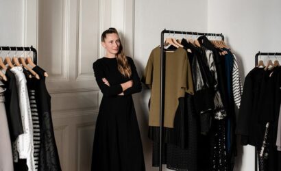 Eine Frau in schwarzem Kleid steht mit verschränkten Armen zwischen zwei Kleiderständern mit hochwertiger, überwiegend dunkler Mode. Sie lächelt leicht und befindet sich in einem stilvollen, hellen Raum mit klassischer Wandvertäfelung.