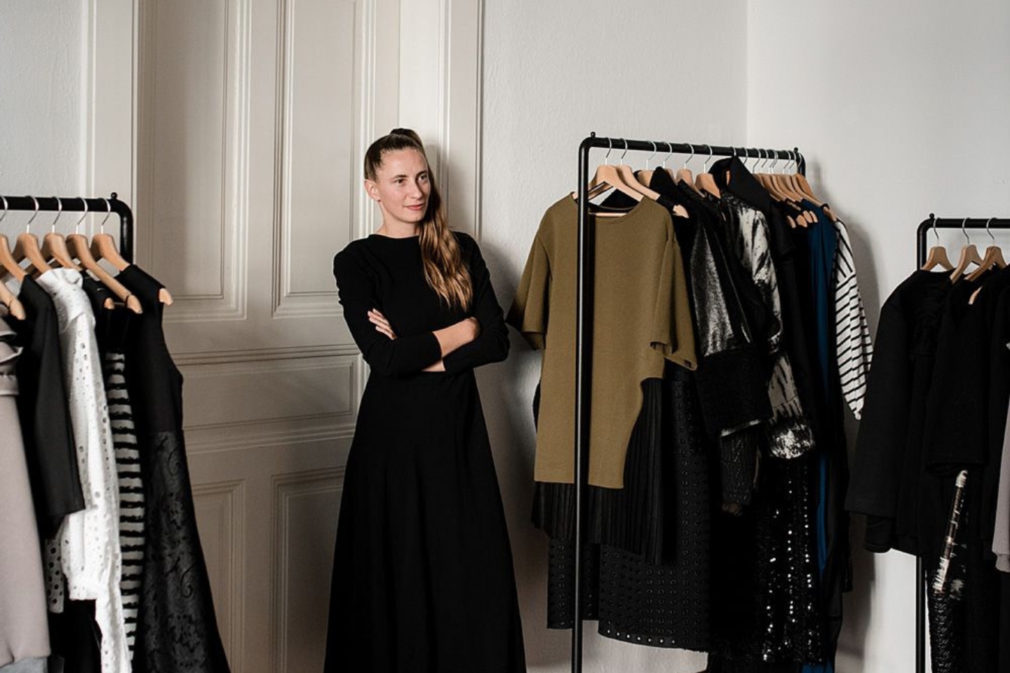 Eine Frau in schwarzem Kleid steht mit verschränkten Armen zwischen zwei Kleiderständern mit hochwertiger, überwiegend dunkler Mode. Sie lächelt leicht und befindet sich in einem stilvollen, hellen Raum mit klassischer Wandvertäfelung.