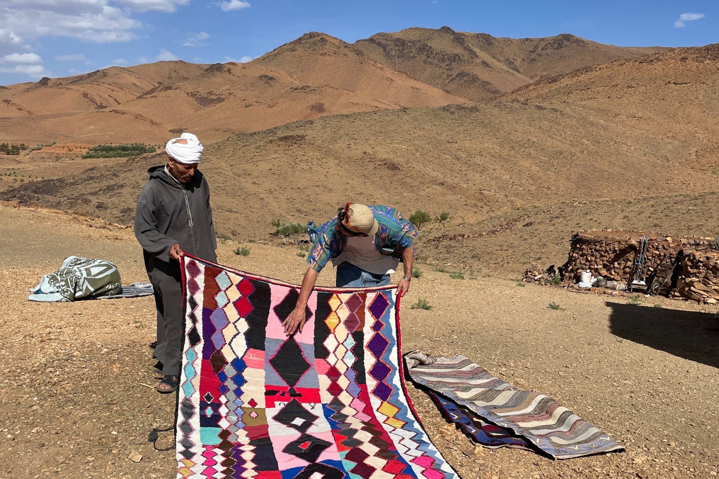 Zwei Männer stehen in einer trockenen, bergigen Landschaft und halten gemeinsam einen bunten, handgewebten Teppich. Einer der Männer trägt traditionelle Kleidung mit weißem Turban, der andere ein buntes Hemd und eine Kappe. Um sie herum liegen weitere Teppiche auf dem Boden ausgebreitet. Im Hintergrund sind Berge, ein paar Pflanzen und eine kleine Steinhütte zu sehen.