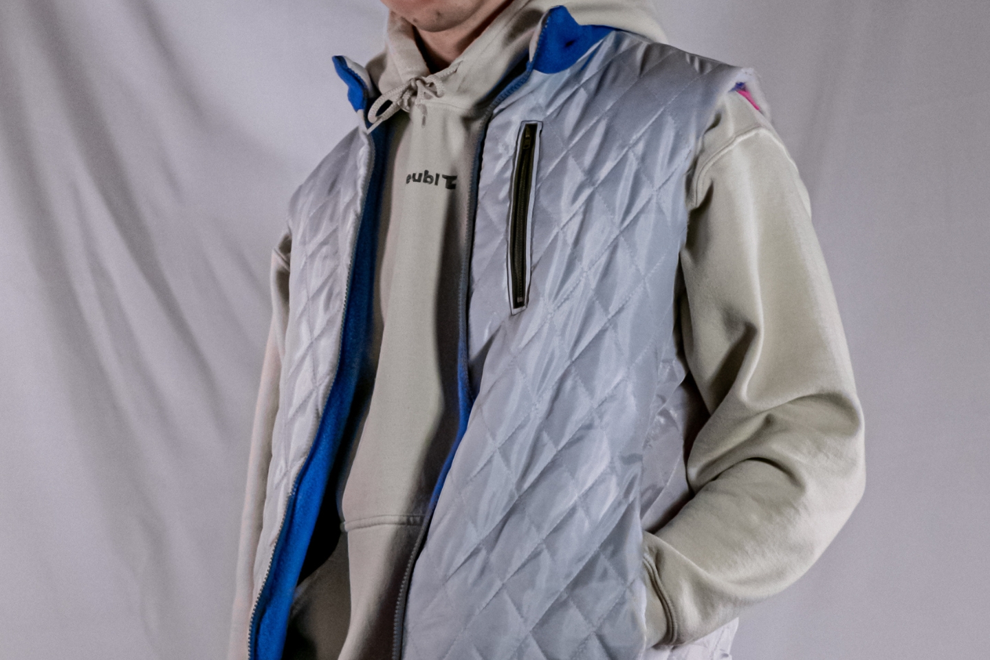 Eine Person trägt eine gesteppt reflektierende silbergraue Weste mit Reißverschluss über einem cremefarbenen Kapuzenpullover. Die Weste hat blaue Innenseiten und eine Reißverschlusstasche auf der Brust. Der Hintergrund ist neutral grau und lässt das Outfit im Fokus stehen.