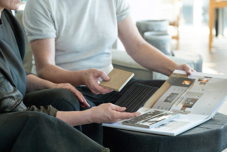 Zwei Personen sitzen nebeneinander und betrachten gemeinsam Materialmuster und ein Designbuch mit Innenarchitekturbeispielen. Eine Person hält ein Holz- und ein schwarzes Lamellenmuster, die andere blättert durch ein Portfolio mit Raumfotos. Der Fokus liegt auf den Händen und den Mustern, die Zusammenarbeit und kreative Planung symbolisieren.