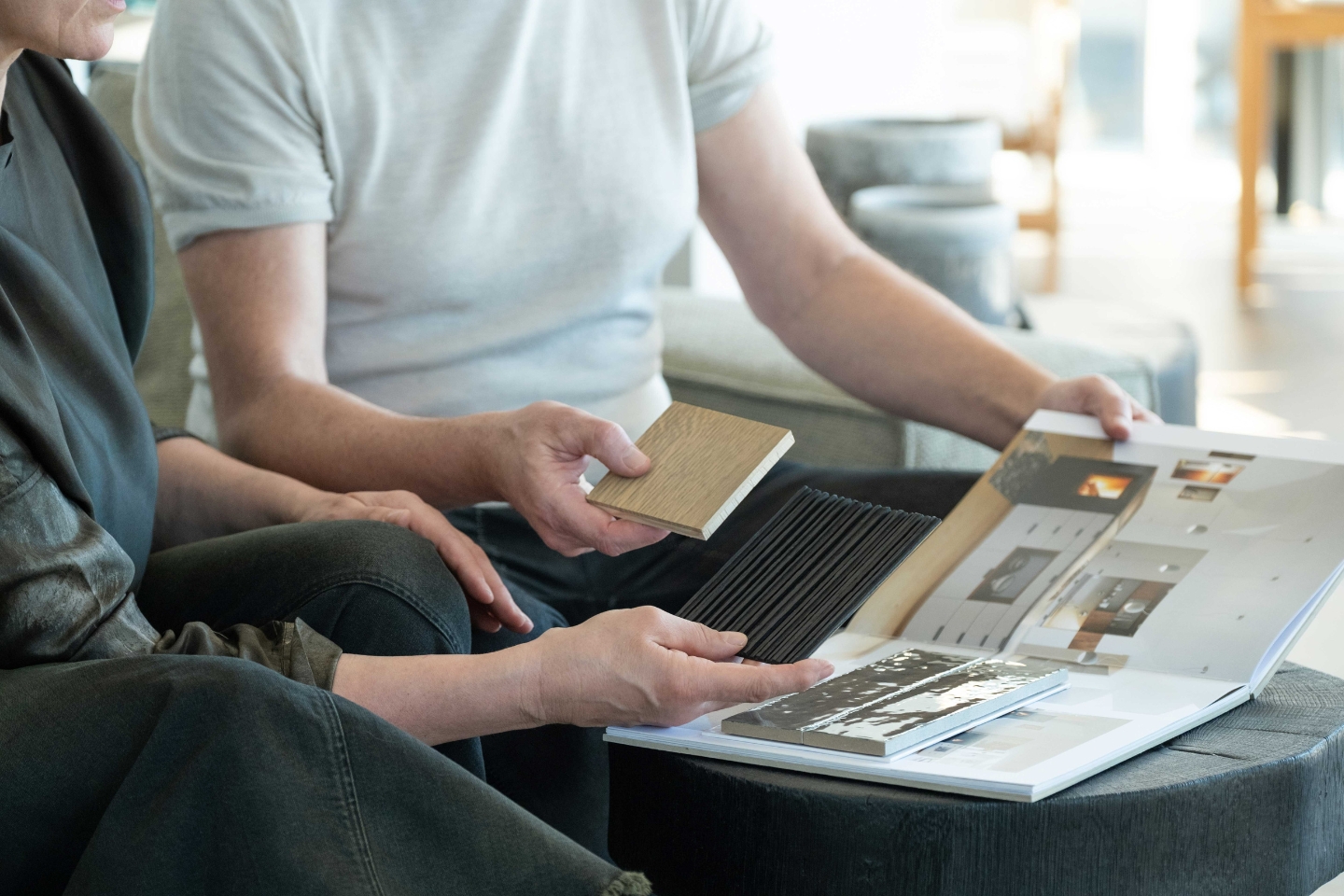 Zwei Personen sitzen nebeneinander und betrachten gemeinsam Materialmuster und ein Designbuch mit Innenarchitekturbeispielen. Eine Person hält ein Holz- und ein schwarzes Lamellenmuster, die andere blättert durch ein Portfolio mit Raumfotos. Der Fokus liegt auf den Händen und den Mustern, die Zusammenarbeit und kreative Planung symbolisieren.
