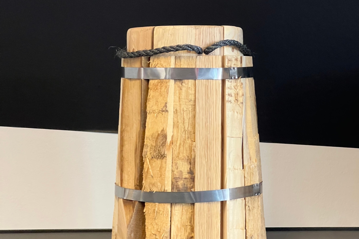 Ein handgefertigter Hocker aus grob bearbeiteten Holzstücken, die vertikal nebeneinander angeordnet und durch zwei metallene Spanngurte zusammengehalten sind. Oben ist eine schwarze Kordel um den Hocker gewickelt. Der Hintergrund besteht aus einer Wand mit schwarzen und weißen Farbflächen.