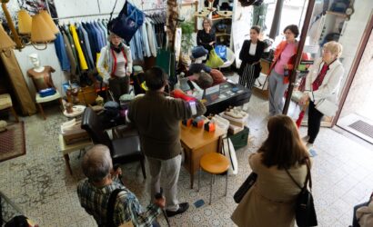 Eine Gruppe von Menschen befindet sich in einem kleinen, dicht eingerichteten Bekleidungsgeschäft. Ein Mann in der Mitte, mit dem Rücken zur Kamera, spricht zur Gruppe. Um ihn herum sind Tische mit Hüten, Schuhkartons, Kleidung an Kleiderstangen, Accessoires und eine Glasvitrine. Die Personen hören aufmerksam zu; manche lehnen an der Wand oder stehen an der Tür. Das Geschäft hat einen gemusterten Fliesenboden und dekorative Lampen.