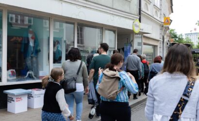 Eine Gruppe von Menschen geht auf einem Gehweg entlang einer Einkaufsstraße. Links im Bild ist ein Schaufenster mit Modeartikeln zu sehen, daneben stehen zwei Hocker mit Aufschriften wie „Creative Industries Styria“, „What now!?“ und „Revolution is now.“ Über der Eingangstür hängt ein Schild mit der Aufschrift „Offline Retail“. Neben der Tür hängt ein Banner mit der Aufschrift „Design in the City“. Die Gruppe scheint an einer geführten Tour teilzunehmen.
