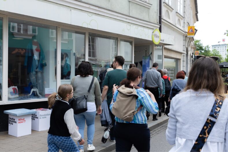 Eine Gruppe von Menschen geht auf einem Gehweg entlang einer Einkaufsstraße. Links im Bild ist ein Schaufenster mit Modeartikeln zu sehen, daneben stehen zwei Hocker mit Aufschriften wie „Creative Industries Styria“, „What now!?“ und „Revolution is now.“ Über der Eingangstür hängt ein Schild mit der Aufschrift „Offline Retail“. Neben der Tür hängt ein Banner mit der Aufschrift „Design in the City“. Die Gruppe scheint an einer geführten Tour teilzunehmen.