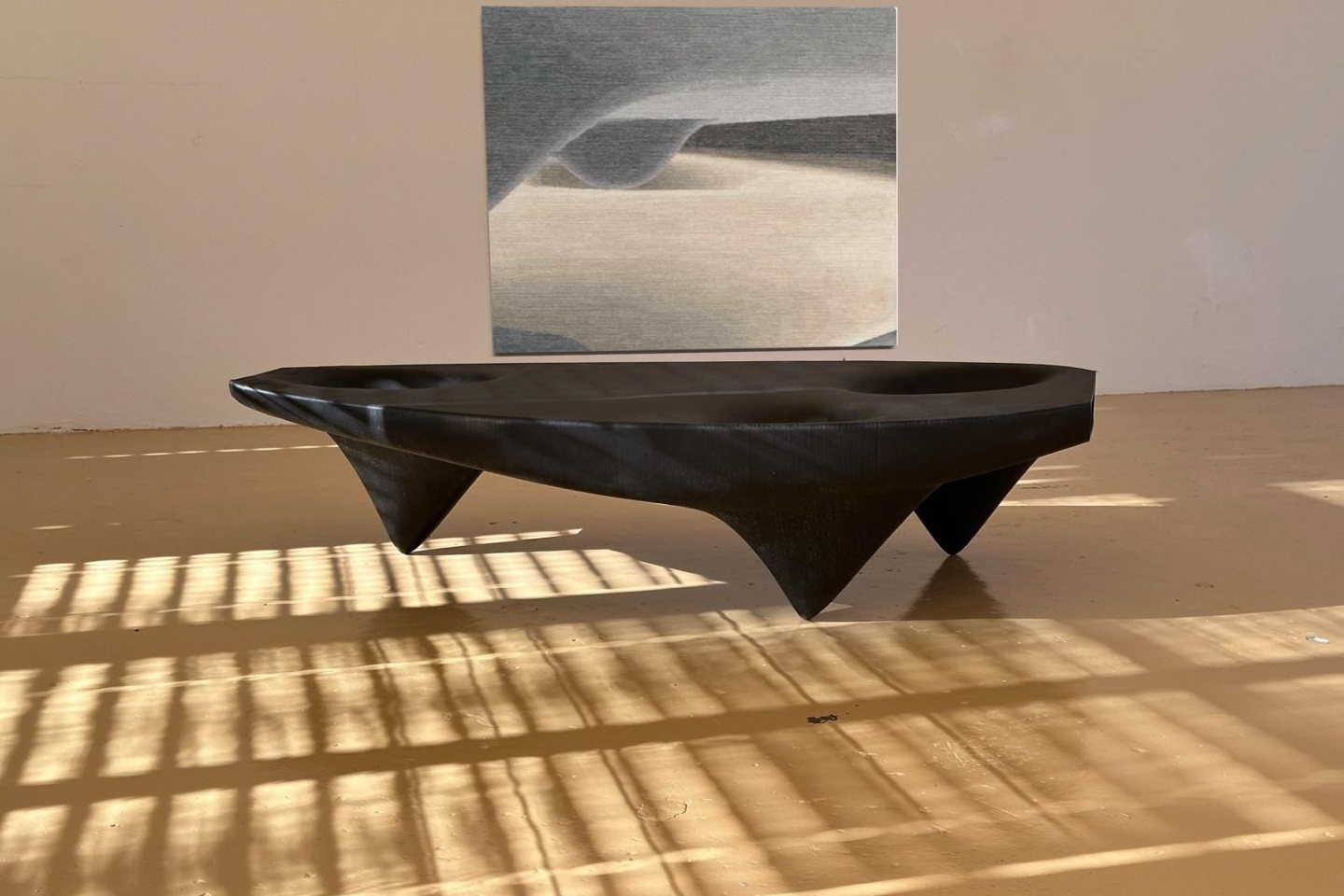 In einem lichtdurchfluteten Raum steht ein skulpturaler, schwarzer Tisch mit geometrischen, spitz zulaufenden Beinen. Hinter dem Tisch hängt ein großformatiges, abstraktes Wandbild in sanften Grau- und Beigetönen, das an eine Landschaft erinnert. Der Boden reflektiert durch Gitterfenster fallendes Sonnenlicht in regelmäßigen Mustern.