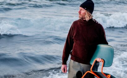 Ein Mann mit blondem, lockigem Haar und Bart steht bis zu den Knien im Meer. Er trägt einen dunkelroten Wollpullover, eine schwarze Mütze und hält einen modernen Stuhl mit orangefarbenem Gestell und grünem Sitz. Der Blick des Mannes geht nach rechts, während hinter ihm Wellen gegen den Strand schlagen. Die Szenerie wirkt rau und kühl.