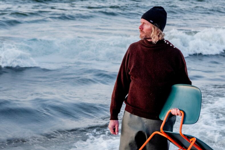 Ein Mann mit blondem, lockigem Haar und Bart steht bis zu den Knien im Meer. Er trägt einen dunkelroten Wollpullover, eine schwarze Mütze und hält einen modernen Stuhl mit orangefarbenem Gestell und grünem Sitz. Der Blick des Mannes geht nach rechts, während hinter ihm Wellen gegen den Strand schlagen. Die Szenerie wirkt rau und kühl.