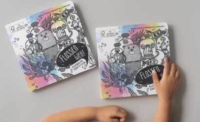 Zwei identische Kinderbücher mit dem Titel „Flossen weg! – Bilderbuch für Kinder & Eltern“ liegen auf einem grauen Tisch. Das Cover zeigt eine bunte, unterwasserartige Illustration mit einem Biber in der Mitte, umgeben von U-Boot-ähnlichen Kreaturen, Pflanzen und Blasen. Die Hintergrundfarben sind in sanften Pastelltönen gehalten. Ein Kinderarm greift nach dem rechten Buch. Oben auf dem Cover steht: „Kinder stärken. Übergriffen vorbeugen.“ Das Buch hat abgerundete Ecken und besteht aus dickem Karton, typisch für Kleinkinderbücher.
