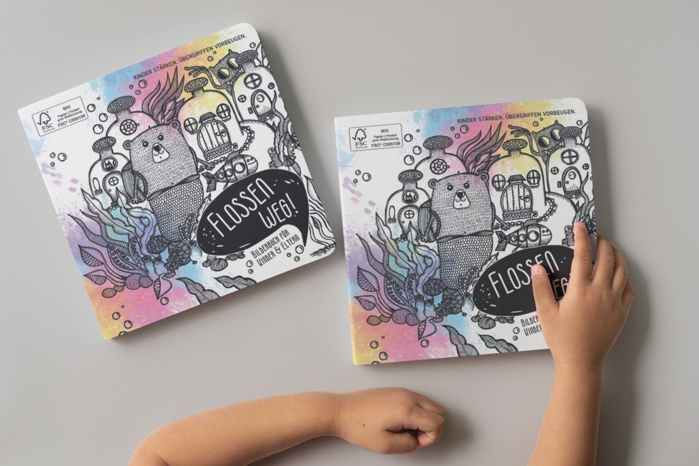 Zwei identische Kinderbücher mit dem Titel „Flossen weg! – Bilderbuch für Kinder & Eltern“ liegen auf einem grauen Tisch. Das Cover zeigt eine bunte, unterwasserartige Illustration mit einem Biber in der Mitte, umgeben von U-Boot-ähnlichen Kreaturen, Pflanzen und Blasen. Die Hintergrundfarben sind in sanften Pastelltönen gehalten. Ein Kinderarm greift nach dem rechten Buch. Oben auf dem Cover steht: „Kinder stärken. Übergriffen vorbeugen.“ Das Buch hat abgerundete Ecken und besteht aus dickem Karton, typisch für Kleinkinderbücher.