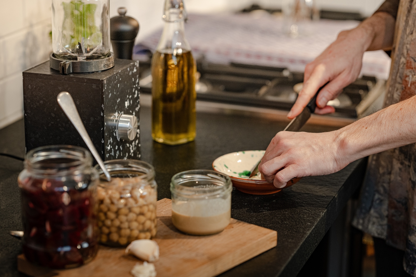 Eine Person bereitet in einer modernen Küche eine Mahlzeit zu. Auf der schwarzen Arbeitsfläche stehen ein schwarzer Mixer mit Glaskrug, ein Holzbrett mit Gläsern voller Kichererbsen, Rote Bete und Tahini sowie eine Knoblauchzehe. Im Hintergrund sind eine Ölflasche und ein Gasherd zu sehen. Die Person hackt Kräuter in einer kleinen Schüssel. Die Szene wirkt gemütlich und gesundheitsbewusst.
