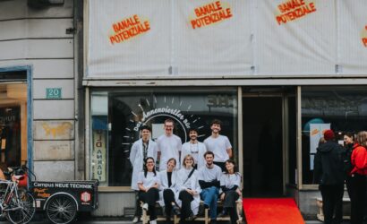 Eine Gruppe junger Menschen in weißen Kitteln und weißen T-Shirts sitzt und steht lächelnd vor einem Schaufenster mit der Aufschrift „Das Annenviertler Büro“. Über dem Fenster ist in roten Buchstaben „BANALE POTENZIALE“ mehrfach gesprüht. Links steht ein Lastenfahrrad mit der Aufschrift „Campus Neue Technik“. Die Stimmung wirkt freundlich und einladend.