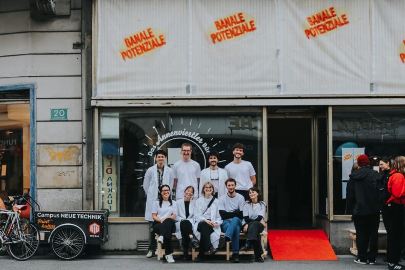 Eine Gruppe junger Menschen in weißen Kitteln und weißen T-Shirts sitzt und steht lächelnd vor einem Schaufenster mit der Aufschrift „Das Annenviertler Büro“. Über dem Fenster ist in roten Buchstaben „BANALE POTENZIALE“ mehrfach gesprüht. Links steht ein Lastenfahrrad mit der Aufschrift „Campus Neue Technik“. Die Stimmung wirkt freundlich und einladend.