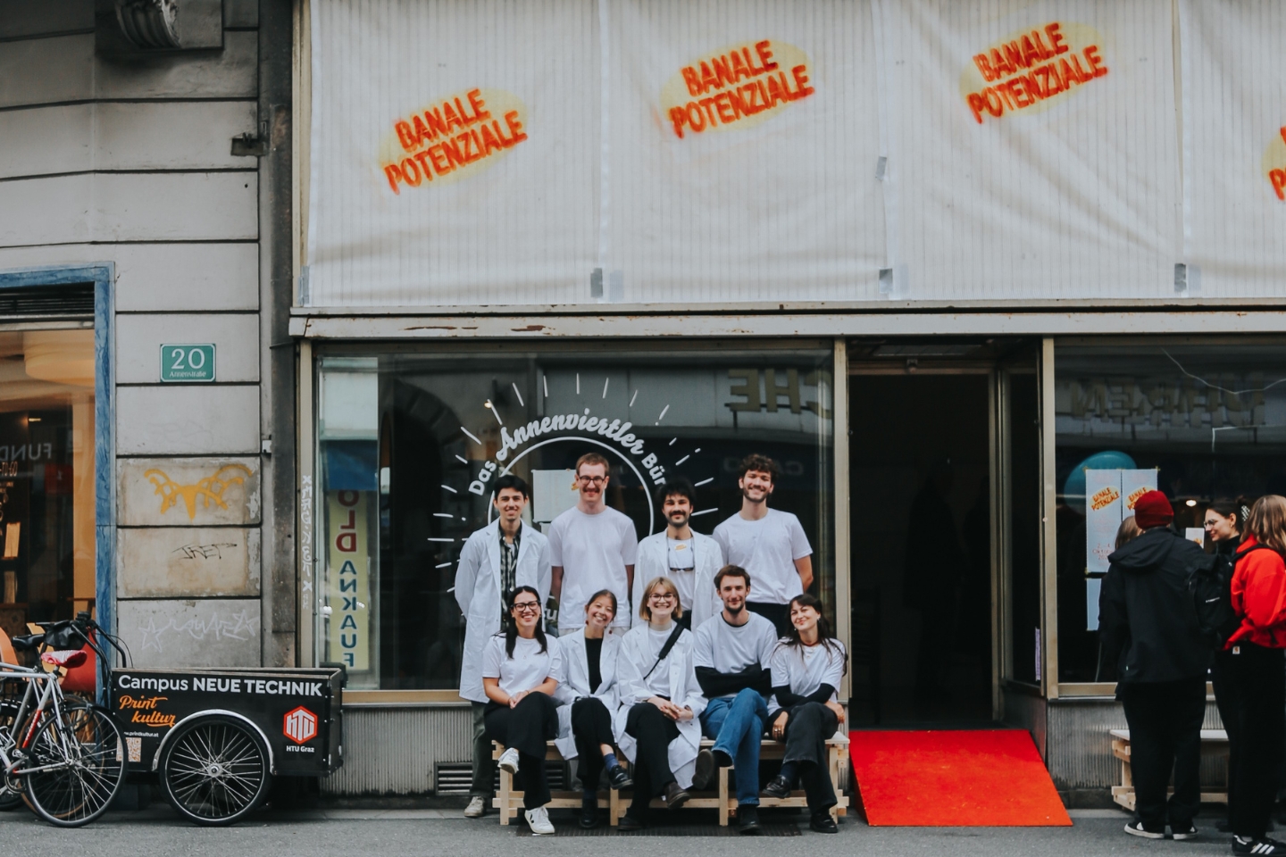 Eine Gruppe junger Menschen in weißen Kitteln und weißen T-Shirts sitzt und steht lächelnd vor einem Schaufenster mit der Aufschrift „Das Annenviertler Büro“. Über dem Fenster ist in roten Buchstaben „BANALE POTENZIALE“ mehrfach gesprüht. Links steht ein Lastenfahrrad mit der Aufschrift „Campus Neue Technik“. Die Stimmung wirkt freundlich und einladend.