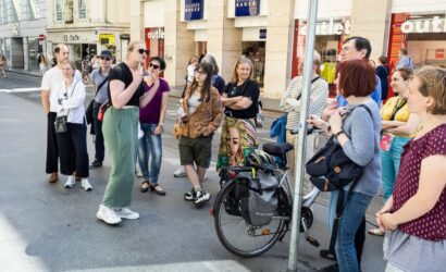 Eine Frau mit Sonnenbrille spricht vor einer Gruppe von Menschen auf einer belebten Einkaufsstraße. Die Gruppe hört aufmerksam zu. Im Hintergrund befinden sich mehrere Geschäfte, darunter ein "Kleider Bauer" und ein "Outlet". Ein Fahrrad mit Gepäcktaschen lehnt an einem Laternenmast im Vordergrund. Die Zuhörenden wirken interessiert und sommerlich gekleidet.
