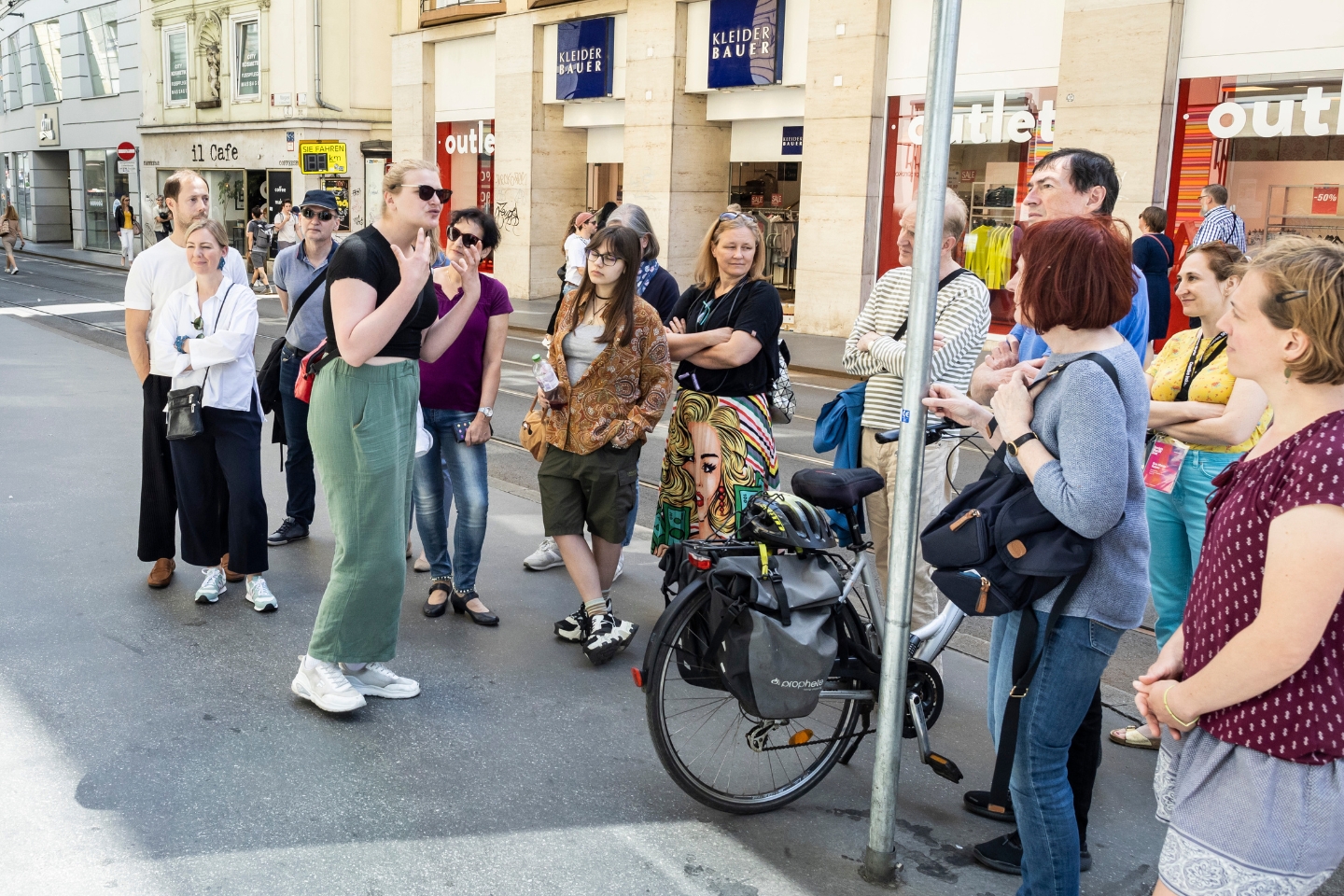 Eine Frau mit Sonnenbrille spricht vor einer Gruppe von Menschen auf einer belebten Einkaufsstraße. Die Gruppe hört aufmerksam zu. Im Hintergrund befinden sich mehrere Geschäfte, darunter ein "Kleider Bauer" und ein "Outlet". Ein Fahrrad mit Gepäcktaschen lehnt an einem Laternenmast im Vordergrund. Die Zuhörenden wirken interessiert und sommerlich gekleidet.