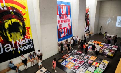 In einer großen Ausstellungshalle mit Betonwänden betrachten zahlreiche Besucher:innen bunte, politische und künstlerische Plakate, die auf dem Boden ausgelegt und an den Wänden aufgehängt sind. Die Banner zeigen markante Texte wie „Ja! Nein! Nein!“, „DEMO CRAZY“ und „Take a walk on the wild side“.