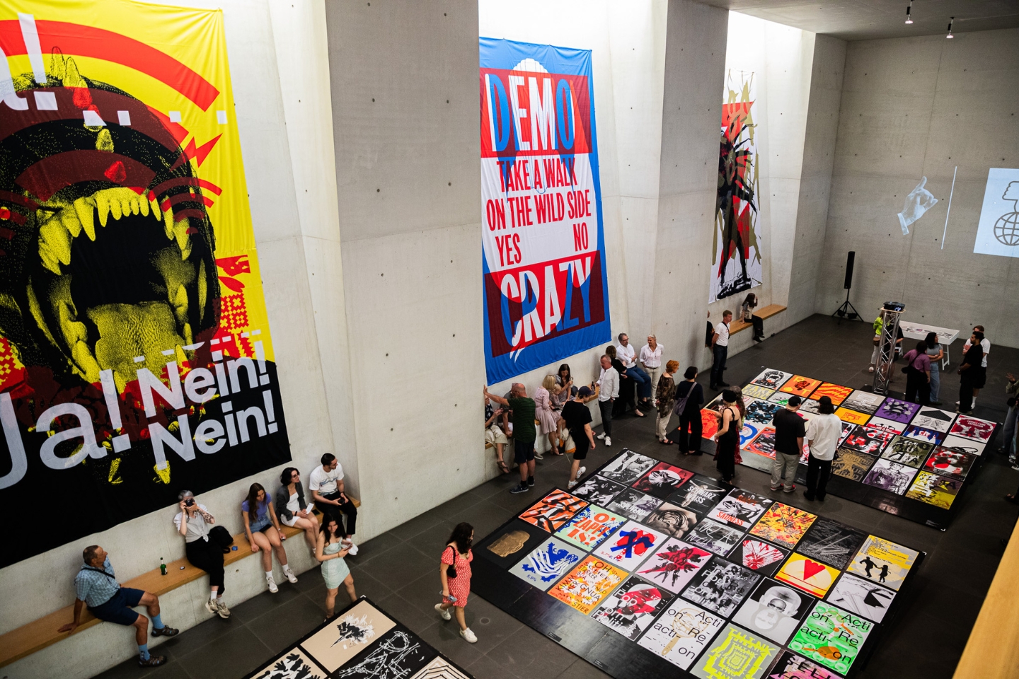 In einer großen Ausstellungshalle mit Betonwänden betrachten zahlreiche Besucher:innen bunte, politische und künstlerische Plakate, die auf dem Boden ausgelegt und an den Wänden aufgehängt sind. Die Banner zeigen markante Texte wie „Ja! Nein! Nein!“, „DEMO CRAZY“ und „Take a walk on the wild side“.